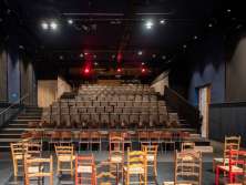 A stage with many different wooden chairs. A seated grandstand. The entrance is on the right.