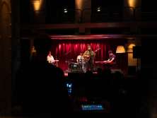 A four-piece band. The lead singer is standing with a guitar. The mixing desk is in the foreground in the center of the picture.