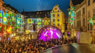 Stars in the city A square between houses. The facades are colorfully illuminated. In the middle is a stage with musicians. The square is filled with people.