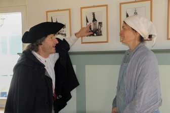 A person in a black cloak and black hat points to a picture with a burning tower on it. A second person in a light-colored dress and white headscarf listens intently.