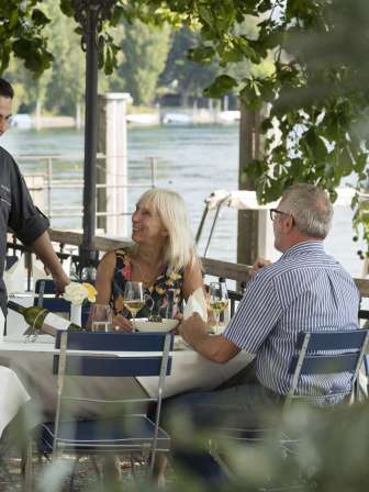 Two people are sitting at a table set in white in the chestnut garden. The chef stands at the table and chats with the guests. The Rhine can be seen in the background.