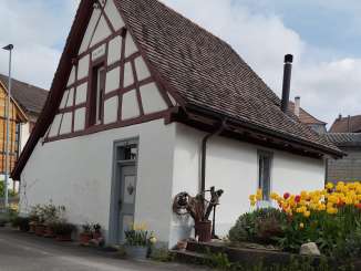 An old small house with a white façade and brown beams. To the right is a garden with various flowers.