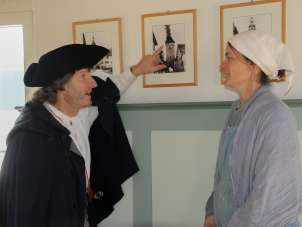 A person in a black cloak and black hat points to a picture with a burning tower on it. A second person in a light-colored dress and white headscarf listens intently.