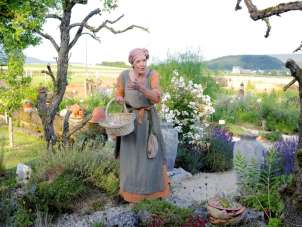 A woman in an organic and green robe with a headscarf stands in a blooming garden. She has a basket in her hand.