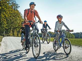 A family cycles on a dirt road, surrounded by meadows, fields, and a forest.
