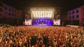 Many people with their arms in the air are at a concert with a band on a large stage.
