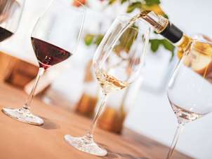 There are four wine glasses on a table. Two of them are filled with red wine, one is empty and white wine is being poured into one.