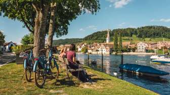 Two friends are sitting on a wooden bench by the Rhine with a view of Stein am Rhein. Behind them are two e-bikes.