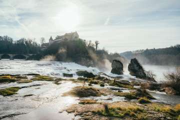 View of the two Rhine Falls rocks and Schloss Laufen from behind. The air is clear in winter and the sun is reflected in the water.