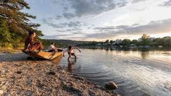 A wooden boat is moored at the shore. Two people are sitting in the boat, enjoying the evening atmosphere. One person is standing in the Rhine, swimming.