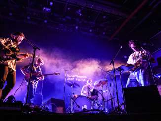 Schaffhausen Jazz Festival A drummer, two electric guitarists and a saxophonist stand on a stage in purple spotlights.