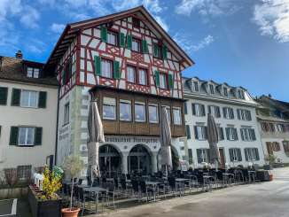 Exterior view of a timber-framed house. There are lots of tables and chairs and three large parasols in front of the house. The house has green shutters and a long wooden bay window.