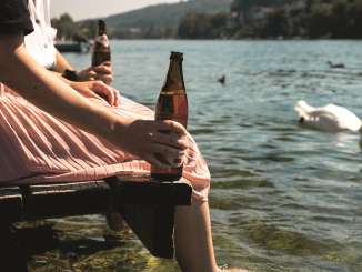 Two friends are sitting on a wooden jetty with their feet dangling in the Rhine. They have a bottle of beer in their hands. A swan swims past.