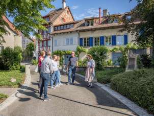 A woman in a traditional costume stands in front of a group and tells stories. The group is standing in an alley with houses with colorful facades.