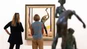Two women are standing in front of a painting depicting a man cutting down a tree with an axe.