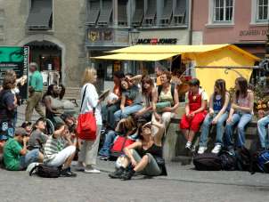 A school class sits on a fountain and square and listens to a guide.