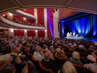 A hall filled with people clapping their hands. An illuminated stage with three people on it, a sofa and an armchair.