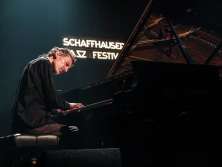 A man sits at a grand piano and plays on a stage. In the background you can see an illuminated sign with the words "Schaffhauser Jazz Festiaval".