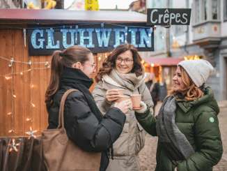 Three friends are wearing thick winter clothing, standing in front of a mulled wine stand and toasting with a mug of mulled wine.