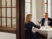 A couple are sitting at a table set in white. They are enjoying a gourmet meal and toasting with a glass of red wine.