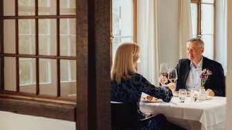 A couple are sitting at a table set in white. They are enjoying a gourmet meal and toasting with a glass of red wine.