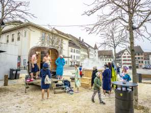 Auf einem grossen Platz steht eine mobile Sauna. Davor stehen Personen im Bademantel.