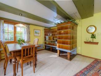 The parlor of a farmhouse has yellow walls, a wooden table with wooden chairs and a tiled stove.