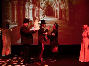 Sechs verkleidete Theaterspielende. Ein grossen Bild von einem Kreuzgang im Hintergrund. Eine Nonne, ein Polizist, ein Herr mit Zilinder, ein Herr mit einer Torte in der Hand.
