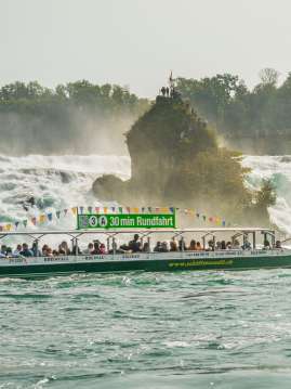 A green boat filled with people. A sign on the boat's roof reads: "30 min Round Trip." The boat is also labeled with www.schiffmaendli.ch. A rock is visible in the background.