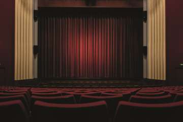 An empty hall with a stage. All the chairs are upholstered in red. The drawn curtain is also red.