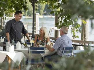 Restaurant Rheinmühle Büsingen Two people are sitting at a table set in white in the chestnut garden. The chef stands at the table and chats with the guests. The Rhine can be seen in the background.