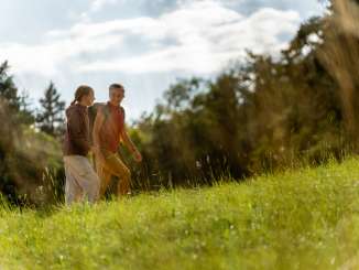 Eine Mutter mit Rucksack wandert mit ihrer Tochter über eine Wiese.