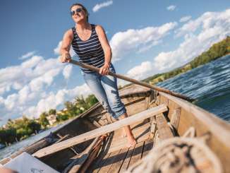 A woman with a striped shirt is standing in the Weidling, a long wooden boat. She is holding an oar in her hand. She uses it to steer the wooden boat.