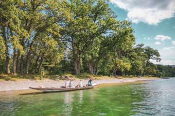Weidling trip Schaffhausen Three people and a dog are sitting in the Weidling, a long wooden boat. A woman is standing at the back, poking along the shore with its pebble beach and trees.