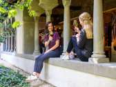 Two friends are sitting on a stone wall in the cloister. Both are holding a glass of wine.