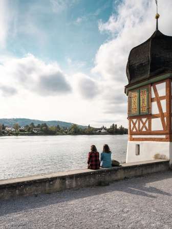 Two friends are sitting on the banks of the Rhine taking a break. To their left is a small bar tower with an onion dome roof.