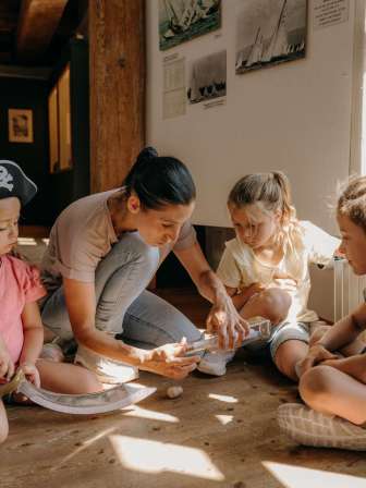 A woman and three children are sitting on the wooden floor. They are opening a message in a bottle. One child is wearing a pirate hat and has a sabre in his hand.