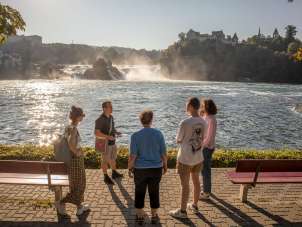 A group of people stand by the Rhine Falls basin. The Rhine Falls can be seen in the background. Everyone is listening intently to one person.