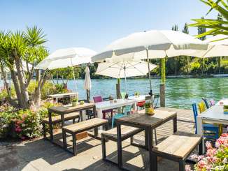 White tables with colorful chairs and white parasols are set up on a wooden jetty on the Rhine.