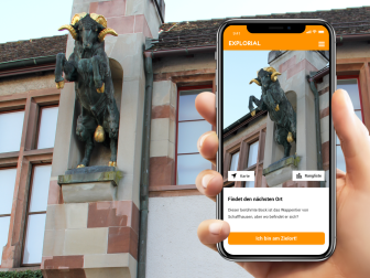 Explorial Schaffhausen A hand holds a cell phone in front of a building with a statue of the Schaffhauser Bock. The picture of the statue can be seen on the cell phone.