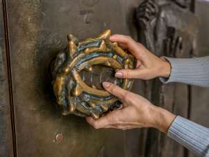 Two hands feel a church door handle. The handle is a rosary with thorns.