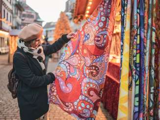 A woman wears a winter coat and beret and looks at a colorful scarf at a Christmas stall.