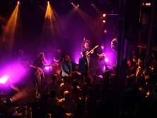 A dark room, an illuminated stage with two singers and five other musicians. The rest of the room is filled with spectators.