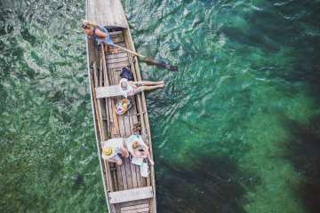 Picture from above of a Weidling, a long and narrow wooden boat. Three people are sitting in the boat and a woman is standing at the back, steering with a wooden rudder. The Rhine shimmers green and blue.