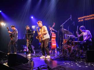 A band with drums, saxophone, double bass, electric guitar and trumpet stands on a stage. Spotlights cast a blue light on the musicians.