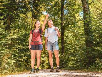 Two friends are standing in the woods, dressed in hiking gear. The woman in the red T-shirt is pointing at something.