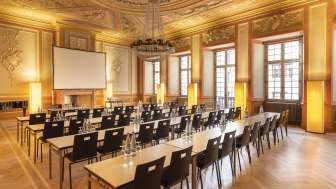 A room with a magnificent stucco ceiling is set up for a seminar. There is a screen in front and rows of tables with chairs behind it.