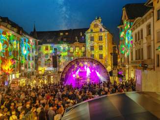 A square between houses. The facades are colorfully illuminated. In the middle is a stage with musicians. The square is filled with people.