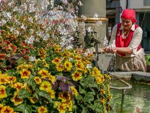 Blooming flowers are visible on the left of the fountain. A lady with a brown skirt and red top holds her hands in the fountain water. She is smiling.