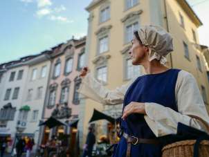The person dressed as a shopkeeper points with her arm to a place of interest.